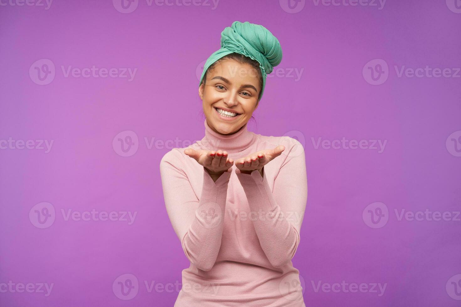 content Jeune magnifique marron aux cheveux femme avec menthe Bandeau en gardant sa paumes élevé tandis que à la recherche à caméra avec charmant sourire, posant plus de violet Contexte photo