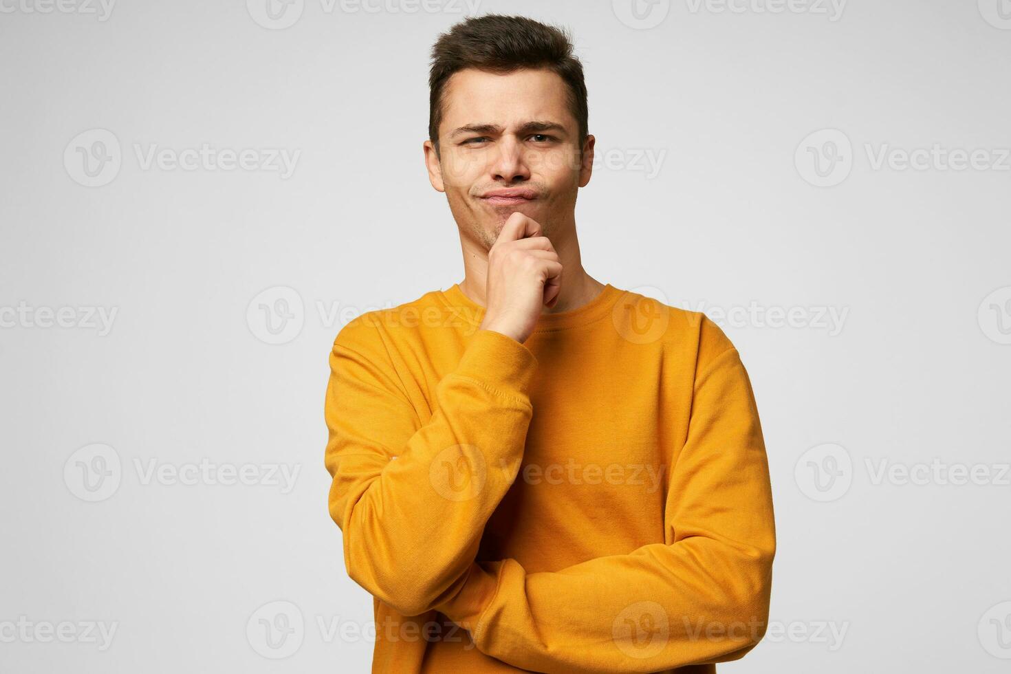 Jeune homme des stands en portant le sien main sur une menton, un coin de le sien lèvres est légèrement pressé, regards pensivement, sérieux, en pensant plus de une problème avec mécontent faciale expression, plus de blanc Contexte photo