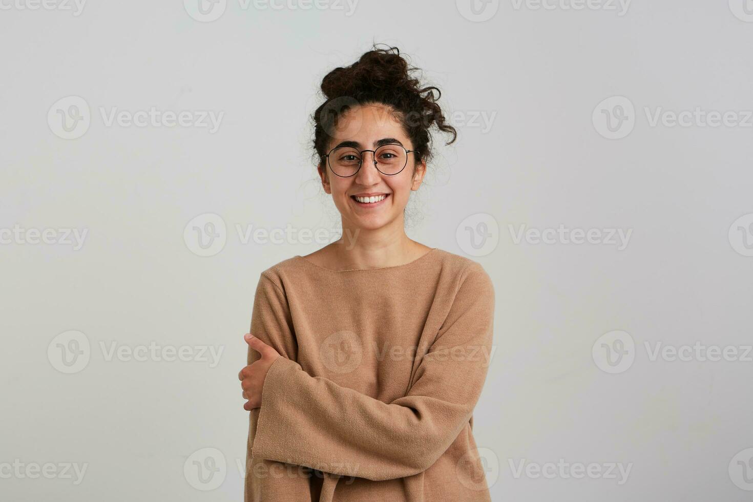 agréable à la recherche femme, magnifique fille avec foncé frisé cheveux chignon. portant beige sauteur et lunettes. émotion concept. détient sa bras et souriant. en train de regarder à le caméra isolé plus de blanc Contexte photo