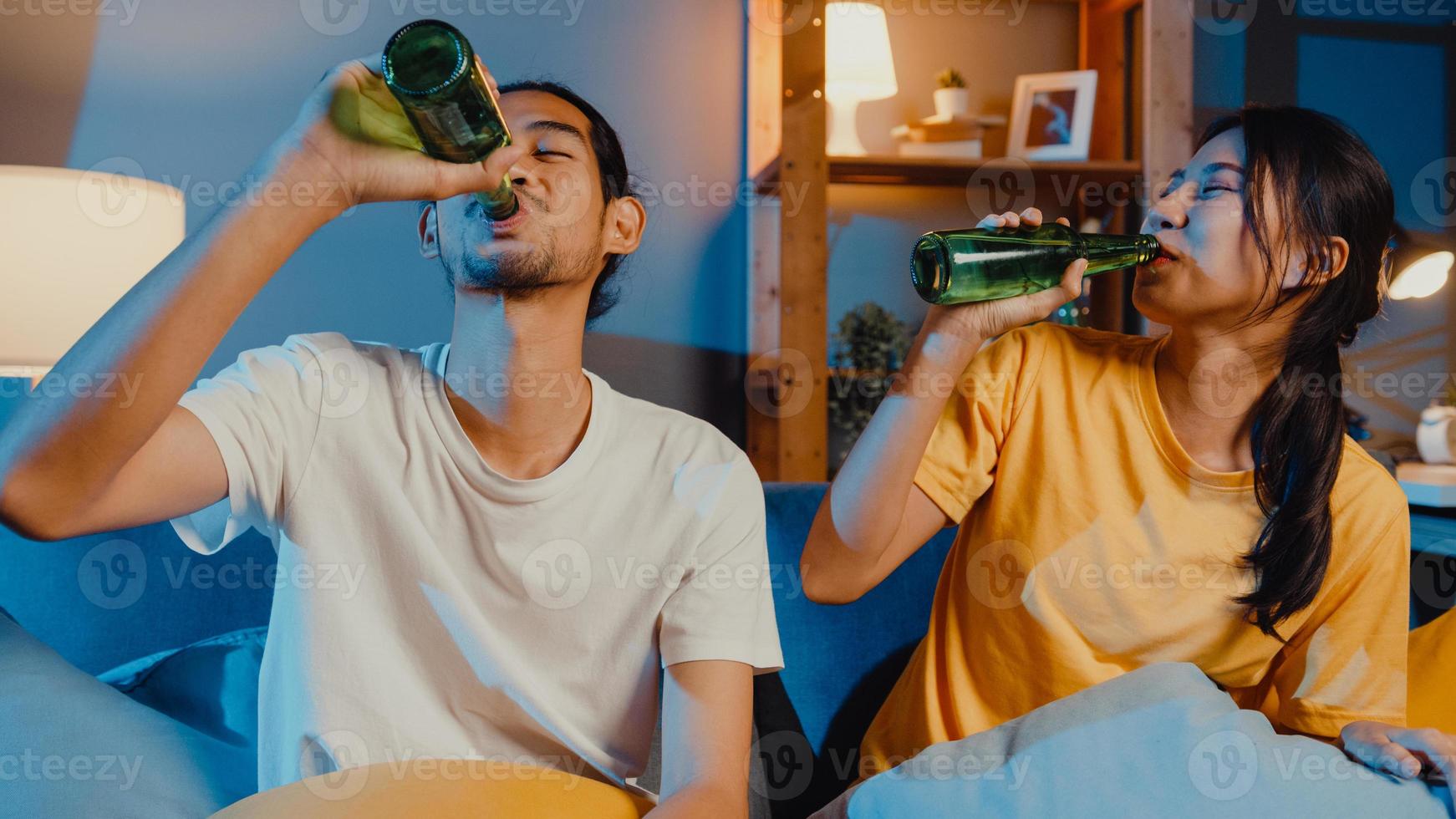 heureux jeune couple asiatique regardant la caméra profiter d'un événement de soirée en ligne assis sur un canapé appel vidéo avec des amis boire de la bière via un appel vidéo en ligne dans le salon à la maison, concept de distanciation sociale. photo