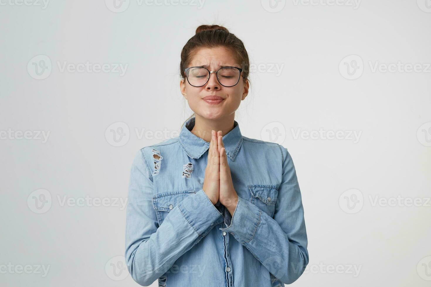 une jolie Jeune fille dans des lunettes des stands avec yeux fermé, mains plié dans de face de son, prier geste, demander pour quelque chose important, vouloir, espérant pour quelque chose isolé sur blanc Contexte photo