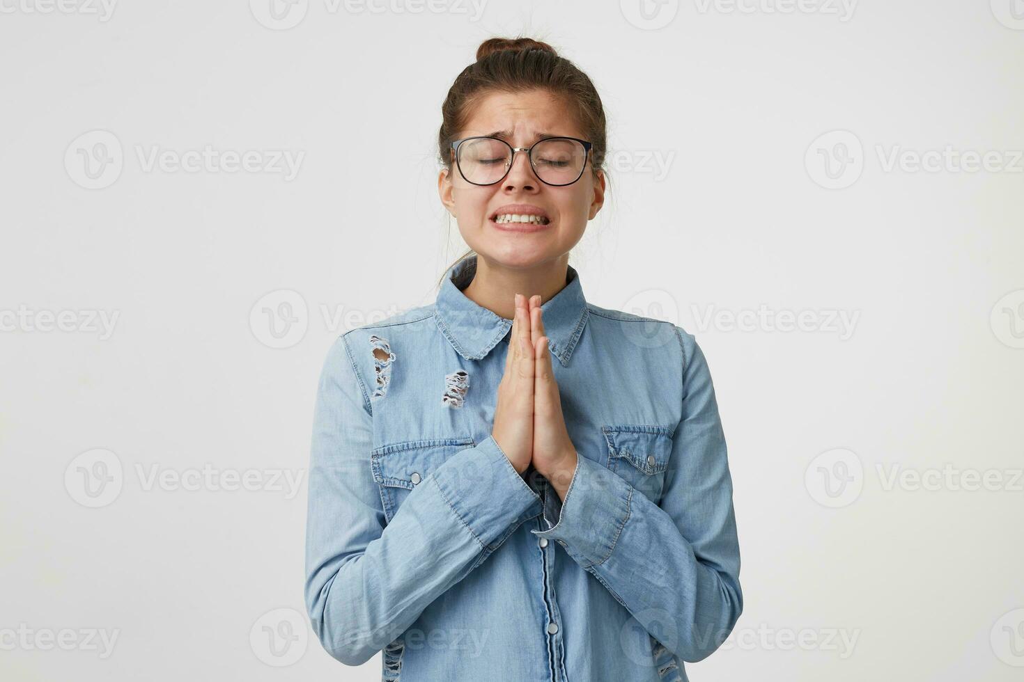 portrait de Jeune fille dans des lunettes des stands avec yeux fermé, mains plié dans de face de son, prier geste, demander pour quelque chose important gravillonnage les dents dur, vouloir, espérant pour quelque chose photo