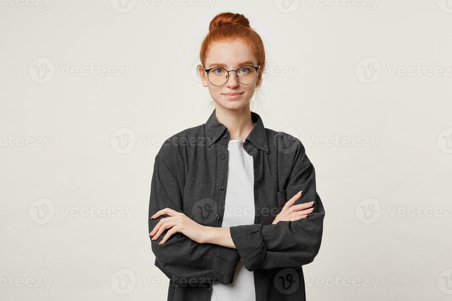 calme confiant roux fille avec cheveux recueillies dans chignon dans une Pour des hommes noir chemise plus de blanc T-shirt, lunettes, des stands avec bras franchi photo