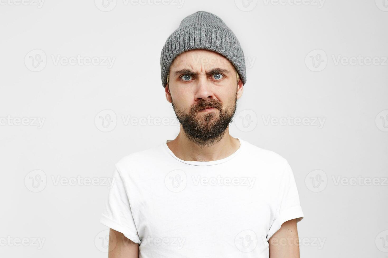 mécontent barbu homme dans une gris chapeau, les sourcils fronça les sourcils, yeux agressif, un coin de le lèvres pincé, habillé dans une Vide blanc T-shirt, isolé plus de blanc Contexte photo