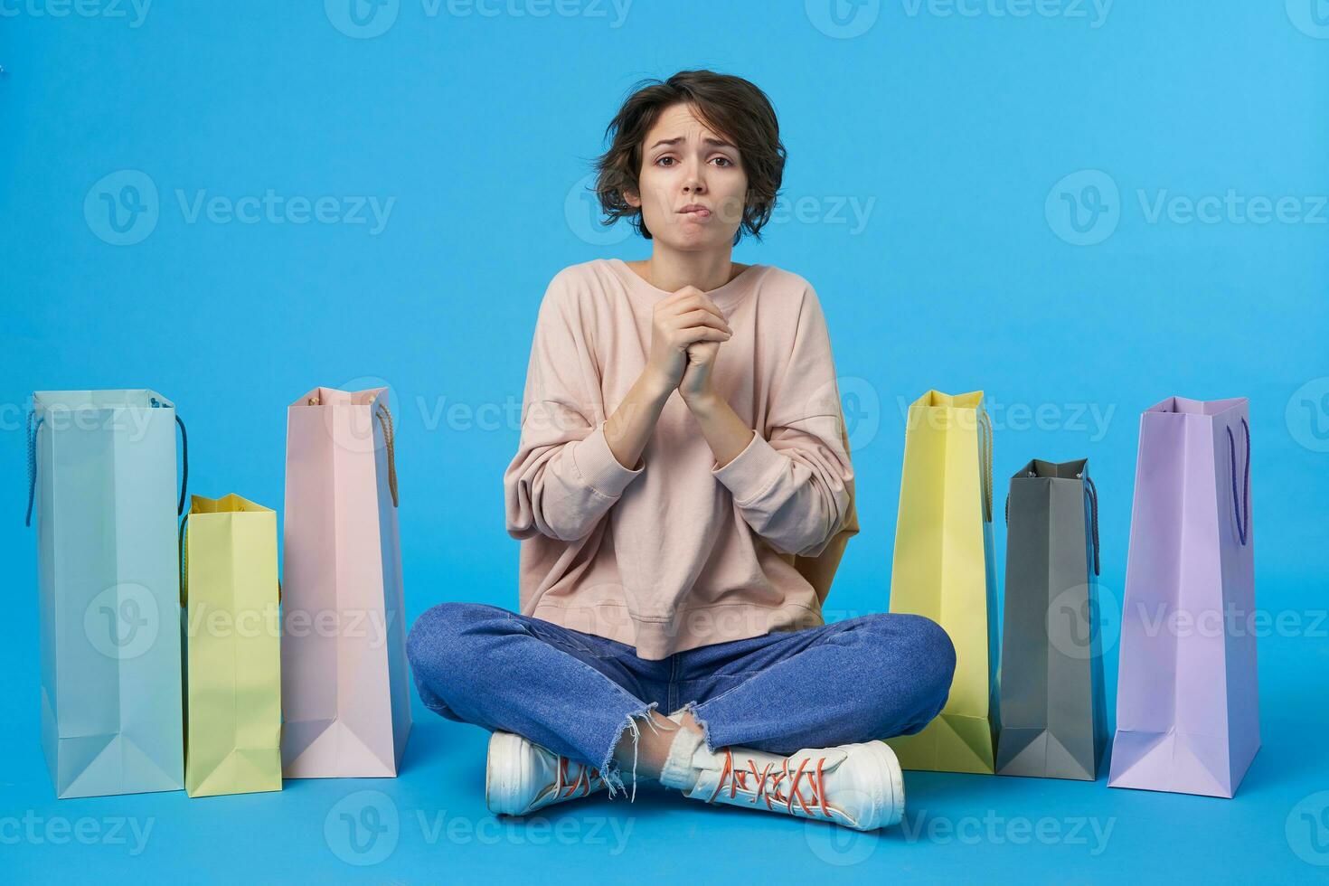 intérieur photo de Jeune foncé aux cheveux frisé Dame à la recherche inquiétant à caméra et pliant élevé mains dans prier geste, posant plus de bleu Contexte avec beaucoup papier Sacs