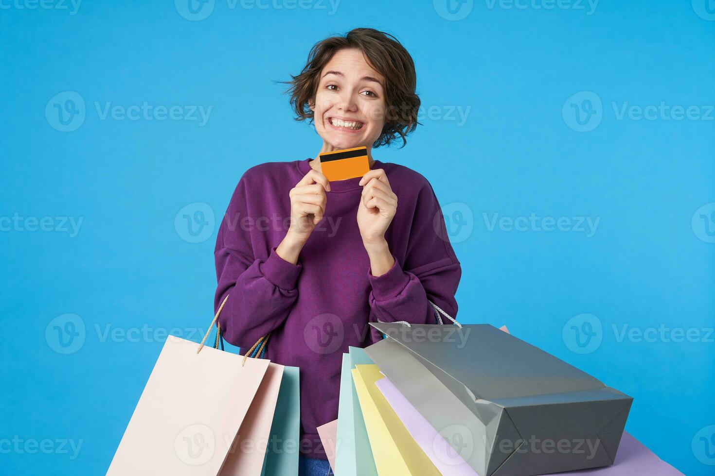 portrait de Jeune content foncé aux cheveux frisé femme montrant sa Plastique carte et souriant largement à caméra, étant dans agréable ambiance tandis que ayant achats jour, isolé plus de bleu Contexte photo