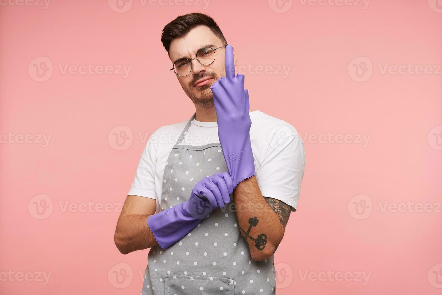 arrogant Jeune court aux cheveux brunette Masculin dans lunettes en mettant sur caoutchouc gants et montrant élevé milieu doigt, portant de base blanc T-shirt et tablier tandis que posant plus de rose Contexte photo