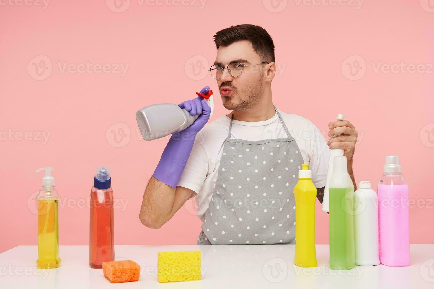 studio photo de Jeune marrant marron aux cheveux Masculin dans des lunettes habillé dans travail vêtements et caoutchouc gants soufflant sur vaporisateur bouteille dans le sien main tandis que imitant pistolet avec il, isolé plus de rose Contexte