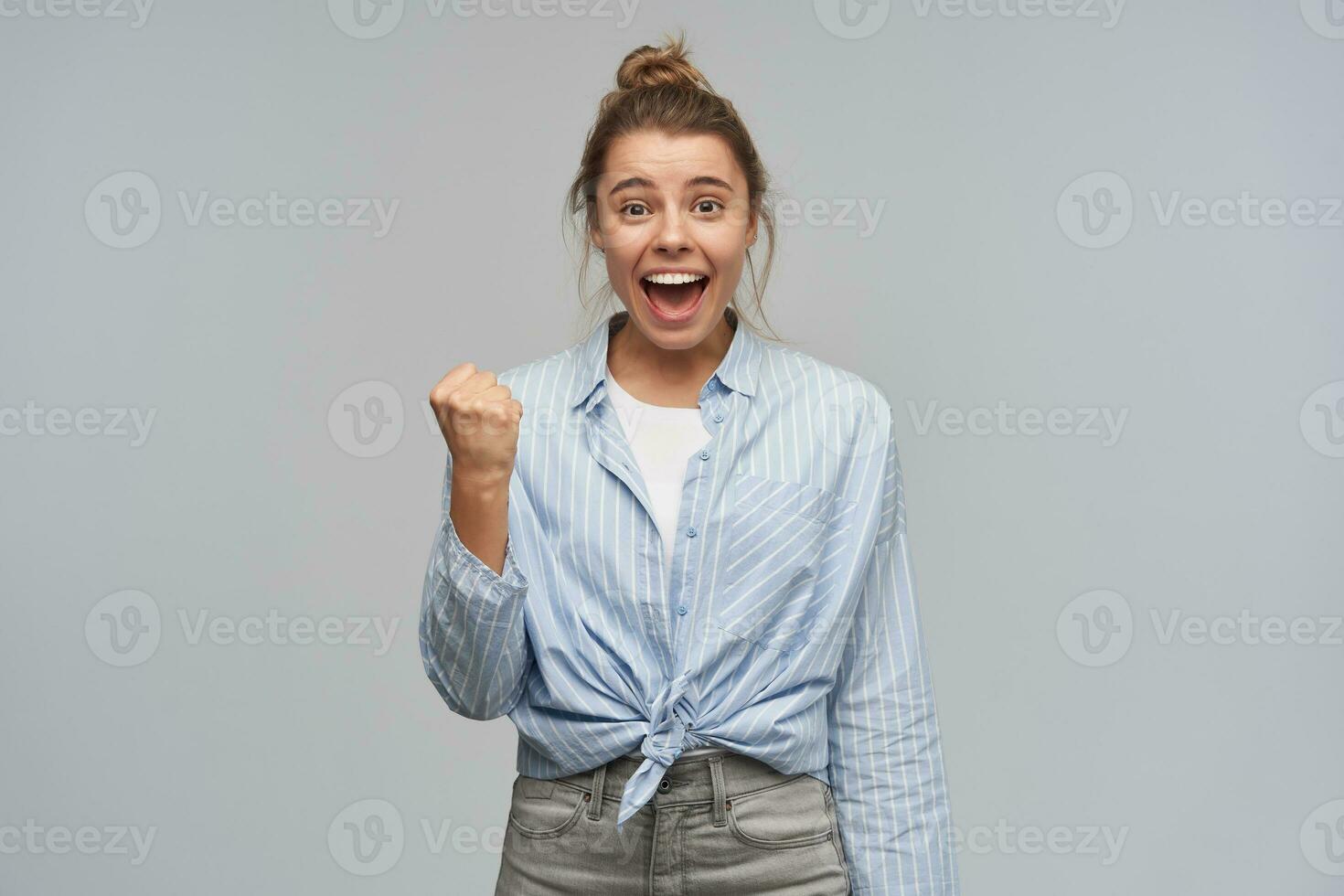 portrait de attractif, excité fille avec blond cheveux recueillies dans chignon. portant rayé noué chemise. serrer sa poing et célébrer succès. en train de regarder à le caméra, isolé plus de gris Contexte photo