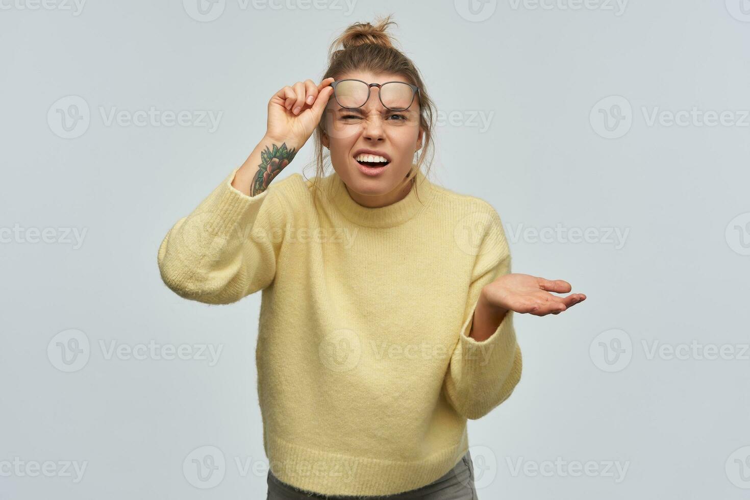 confus femme, débranché fille avec blond cheveux recueillies dans chignon et tatouage. portant Jaune chandail et lunettes. ascenseurs sa des lunettes et hausse les épaules. en train de regarder à le caméra, isolé plus de blanc Contexte photo