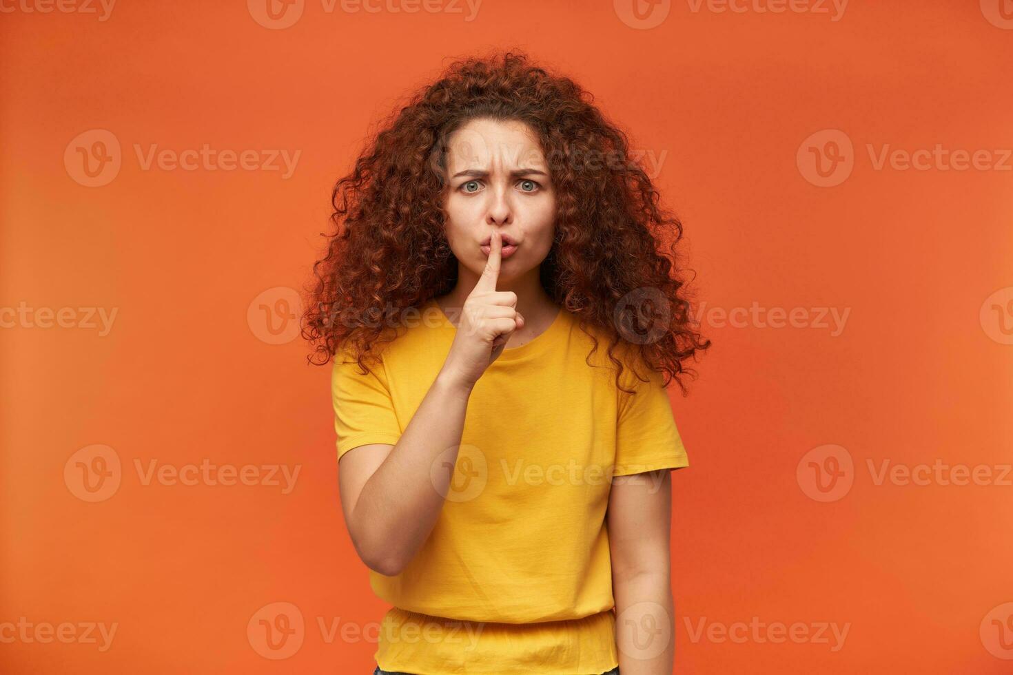 agréable à la recherche femme, magnifique fille avec frisé gingembre cheveux. portant Jaune T-shirt. gens et émotion concept. montrant silence signe et fronce les sourcils. en train de regarder à le caméra isolé plus de Orange Contexte photo