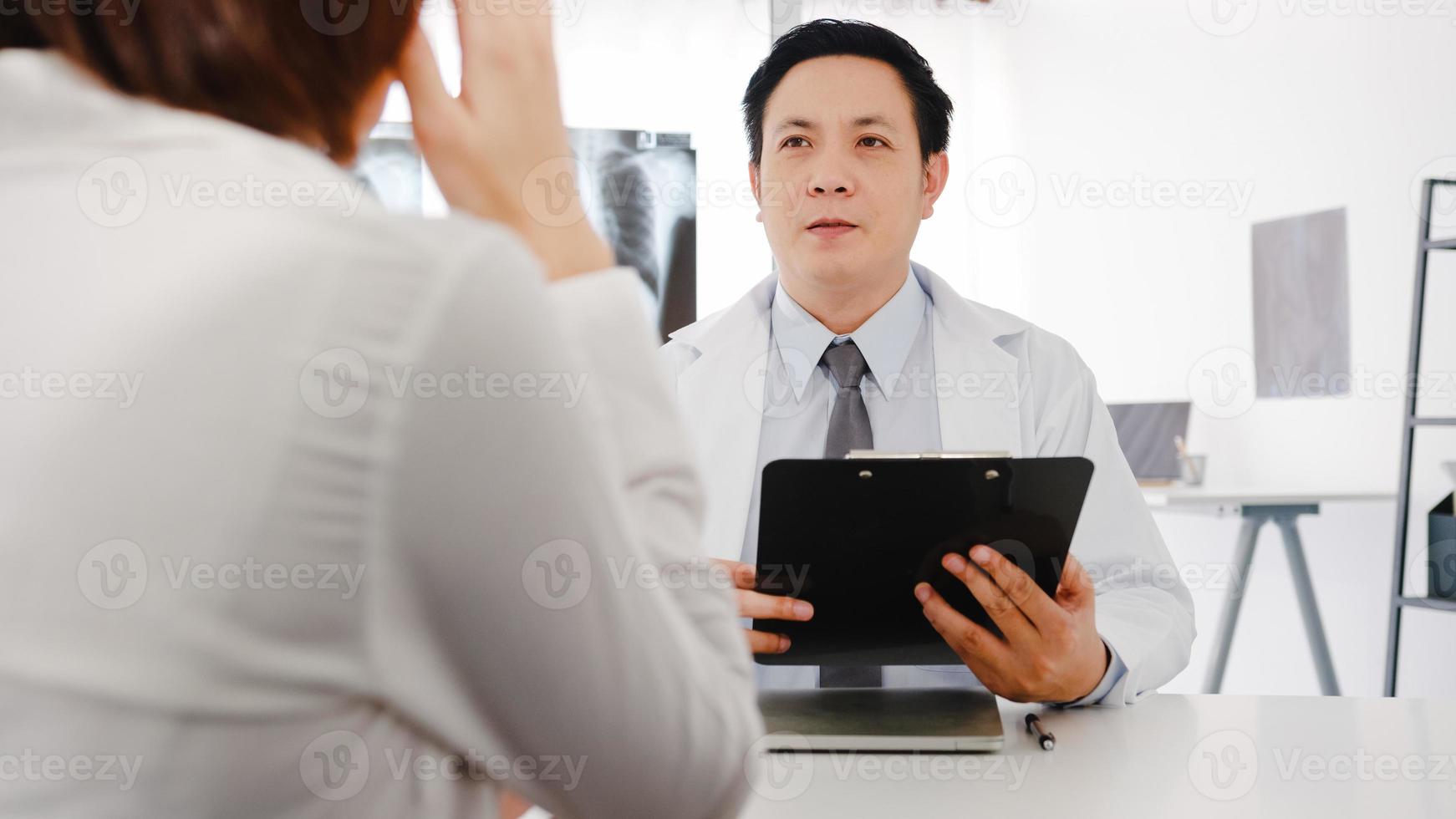 un médecin asiatique sérieux en uniforme médical blanc à l'aide d'un presse-papiers livre d'excellentes nouvelles discutent des résultats ou des symptômes avec une patiente assise au bureau dans une clinique de santé ou un bureau d'hôpital. photo