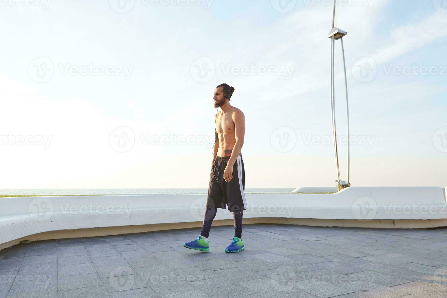 portrait de Jeune sportif homme avec barbe, du repos après jogging, des promenades à le bord de mer, Ecoutez préféré chanson sur écouteurs. jouit grands Matin photo