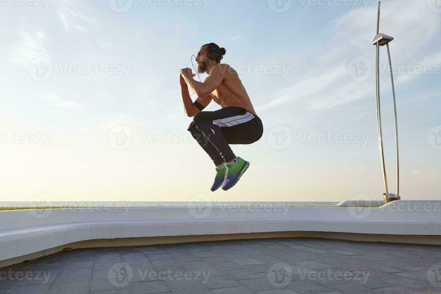 Jeune barbu sauter homme avec en forme athlétique corps, faire Matin des exercices et écoute la musique sur écouteurs, a musclé corps forme. photo