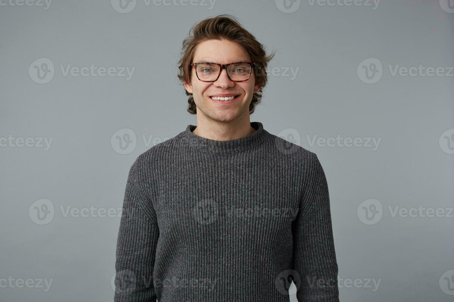 portrait de Jeune de bonne humeur Beau homme avec des lunettes porte dans gris chandail, des stands plus de gris Contexte et largement sourit. photo