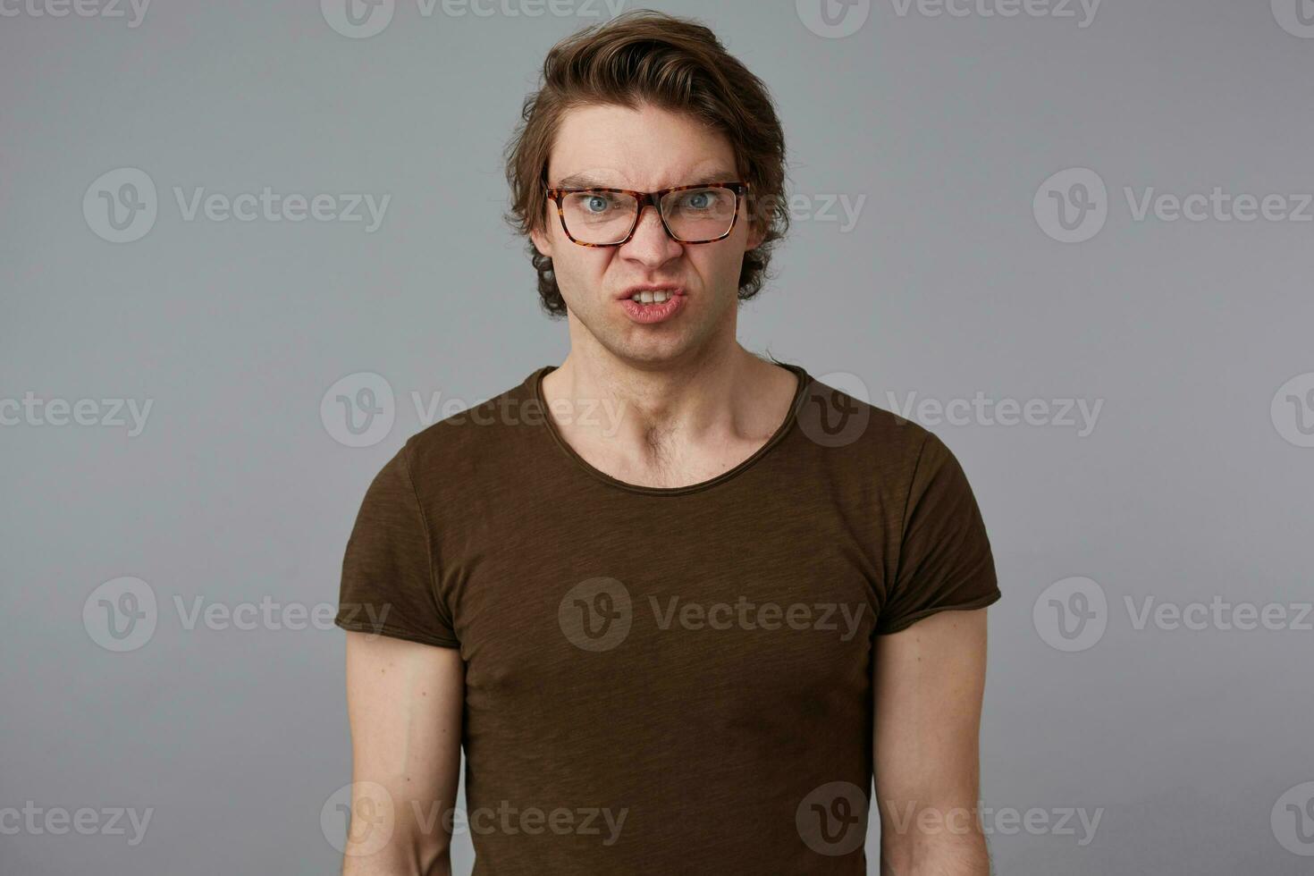 photo de Jeune mécontent gars avec des lunettes porte dans Vide T-shirt, des stands plus de gris Contexte et en colère regards à le caméra.