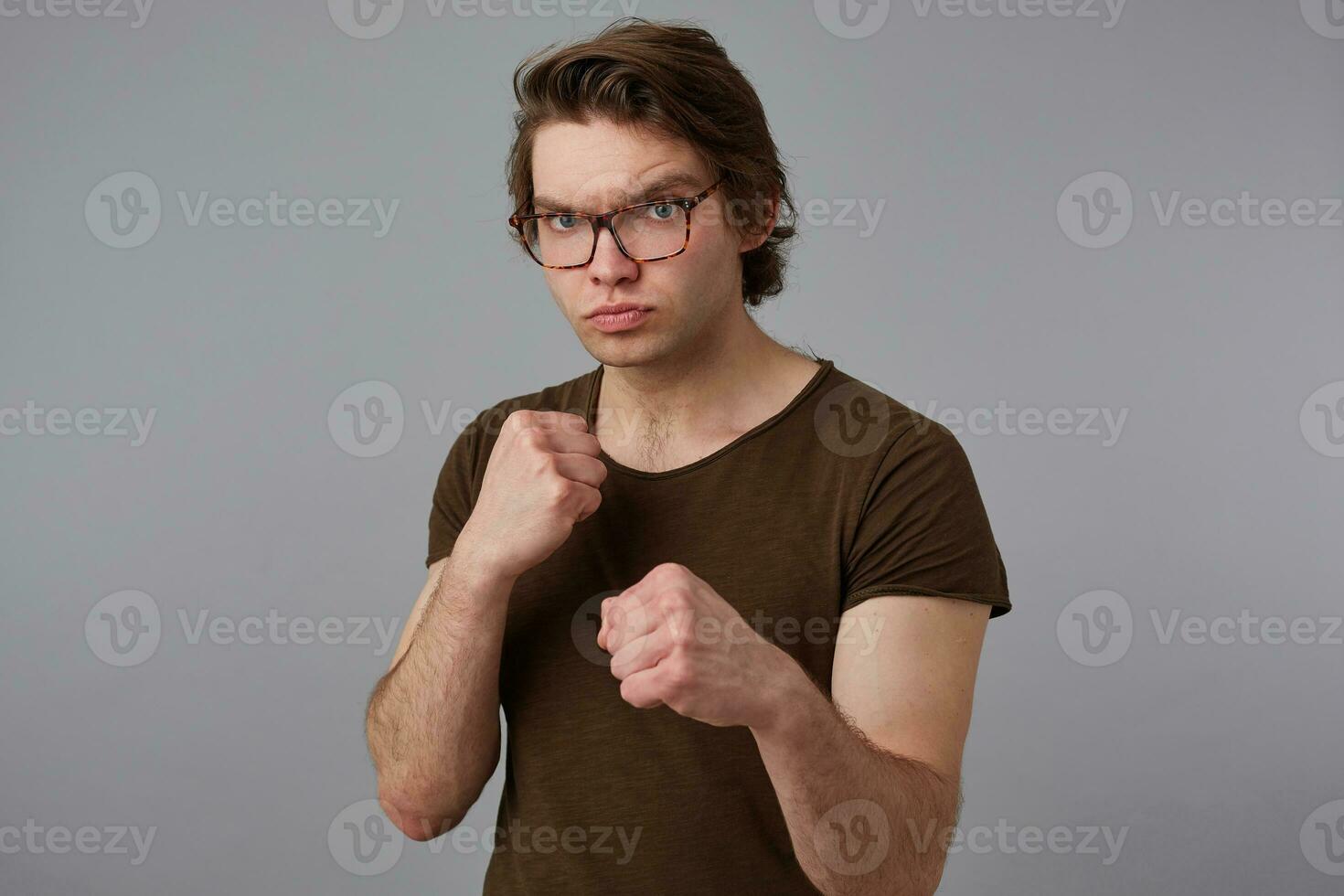 photo de agressif Jeune homme gars avec des lunettes porte dans Vide T-shirt permanent dans défensive posture, en gardant les poings serré, prêt à coup de poing, des stands plus de gris Contexte.