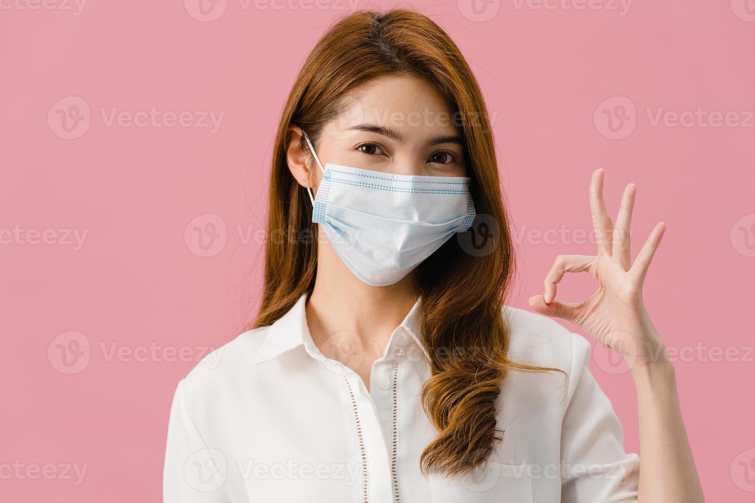 jeune fille asiatique portant un masque médical faisant des gestes signe ok avec vêtue d'un tissu décontracté et regarde la caméra isolée sur fond rose. auto-isolement, distanciation sociale, quarantaine pour le virus corona. photo