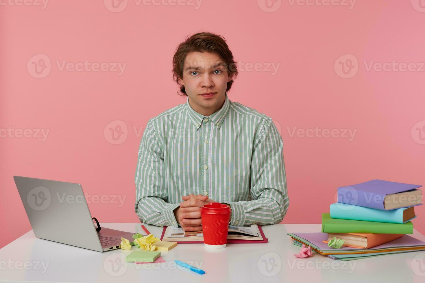 photo de Jeune calme gars avec lunettes, séance à une table avec livres, travail à une ordinateur portable, sérieux regards à le caméra, isolé plus de rose Contexte.