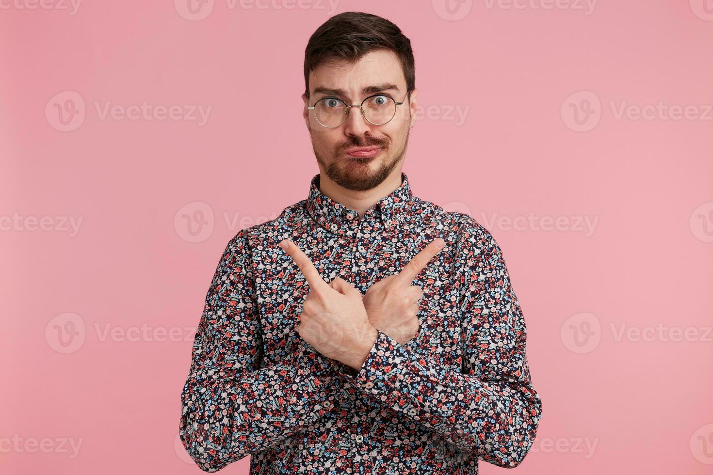 portrait de Jeune homme dans coloré chemise à la recherche pensée des lunettes ayant les doutes, garde mains franchi et montrer du doigt à tous les deux côtés avec indice les doigts isolé plus de rose Contexte. photo