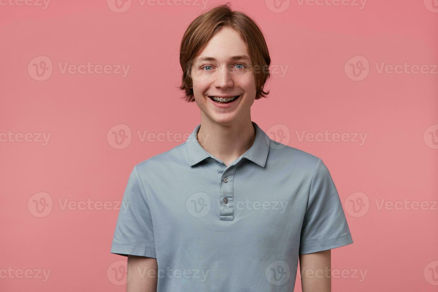 portrait de agréable largement souriant aux yeux bleus soigneusement peigné Jeune gars avec un appareil dentaire sur les dents porte polo T-shirt regards content isolé plus de rose Contexte photo