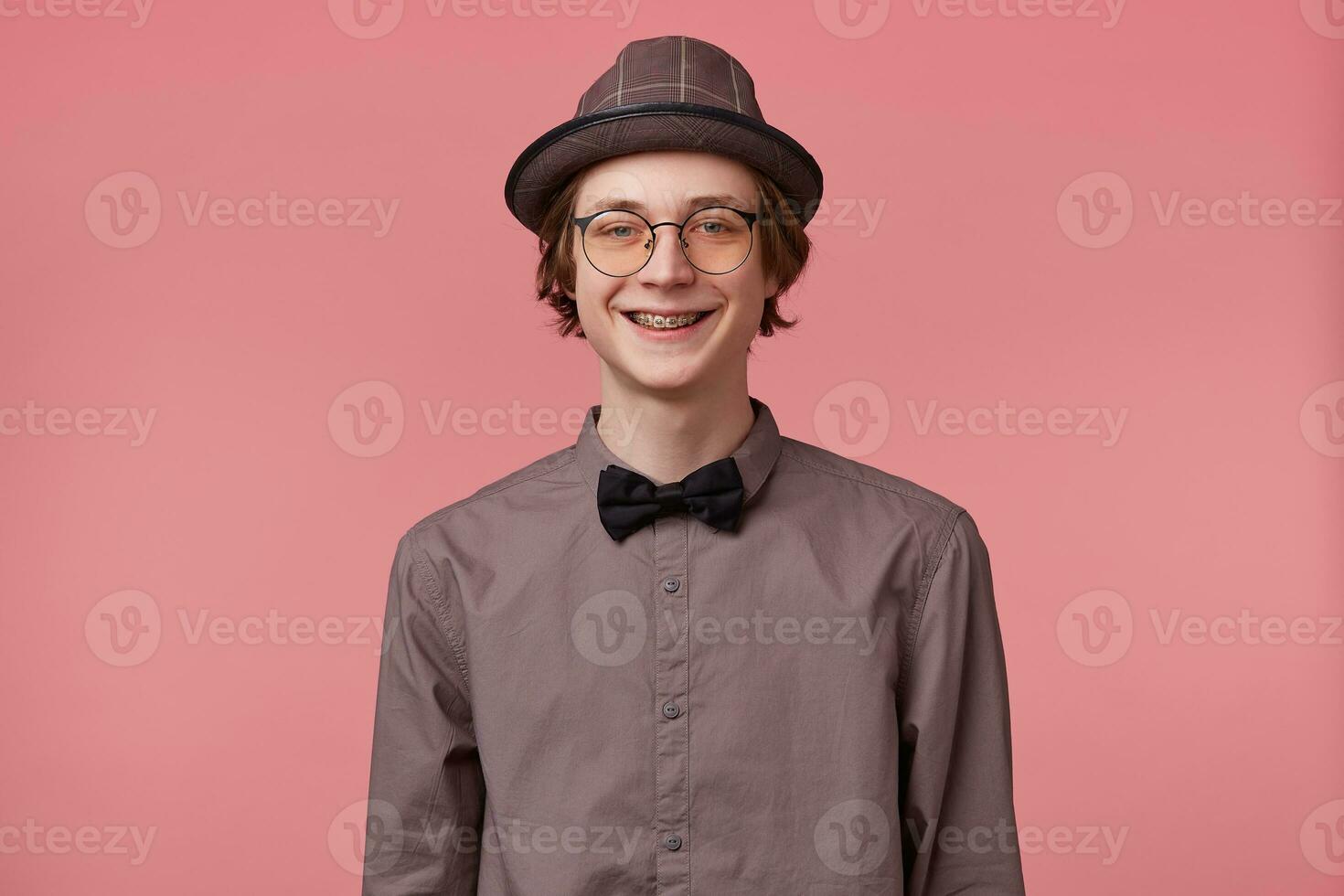 Jeune homme dans chemise chapeau et noir nœud papillon porte des lunettes agréable largement souriant montrant orthodontique supports isolé sur rose Contexte photo