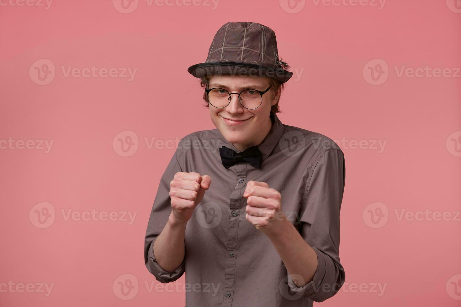 une Jeune mince gars élégamment habillé avec une chapeau des lunettes dans une chemise et arc attacher met vers l'avant le sien les poings comme si Aller à bats toi menace Quelqu'un isolé sur une rose Contexte. photo