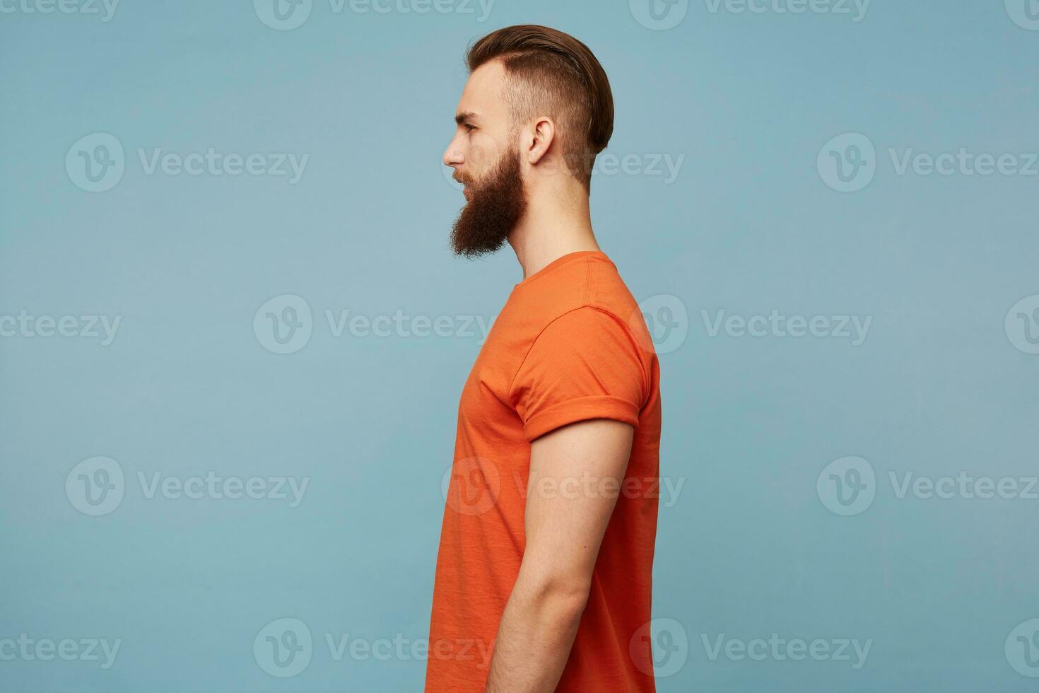 portrait dans profil de une gars avec une à la mode coiffure et une rasé tête et une longue épais barbe dans une rouge T-shirt permanent de côté isolé sur une bleu Contexte photo