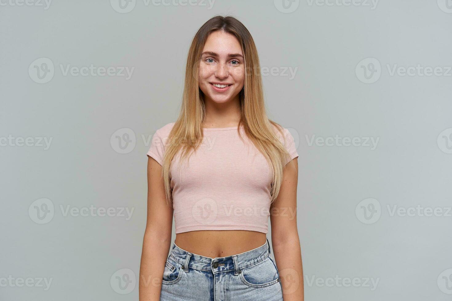 adolescent fille, content à la recherche femme avec blond longue cheveux. portant rose T-shirt et jeans. gens et émotion concept. en train de regarder avec sourire à le caméra isolé plus de gris Contexte photo