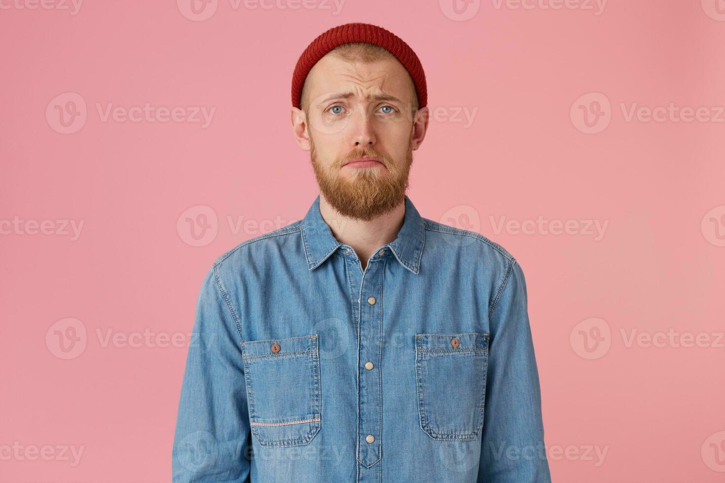 portrait de aux yeux bleus Jeune gars dans rouge chapeau avec rouge barbe regards triste, bouleversé, frustré, mécontent avec quelque chose, lèvres moue, exprime insulte, porte à la mode denim chemise, isolée sur rose Contexte photo