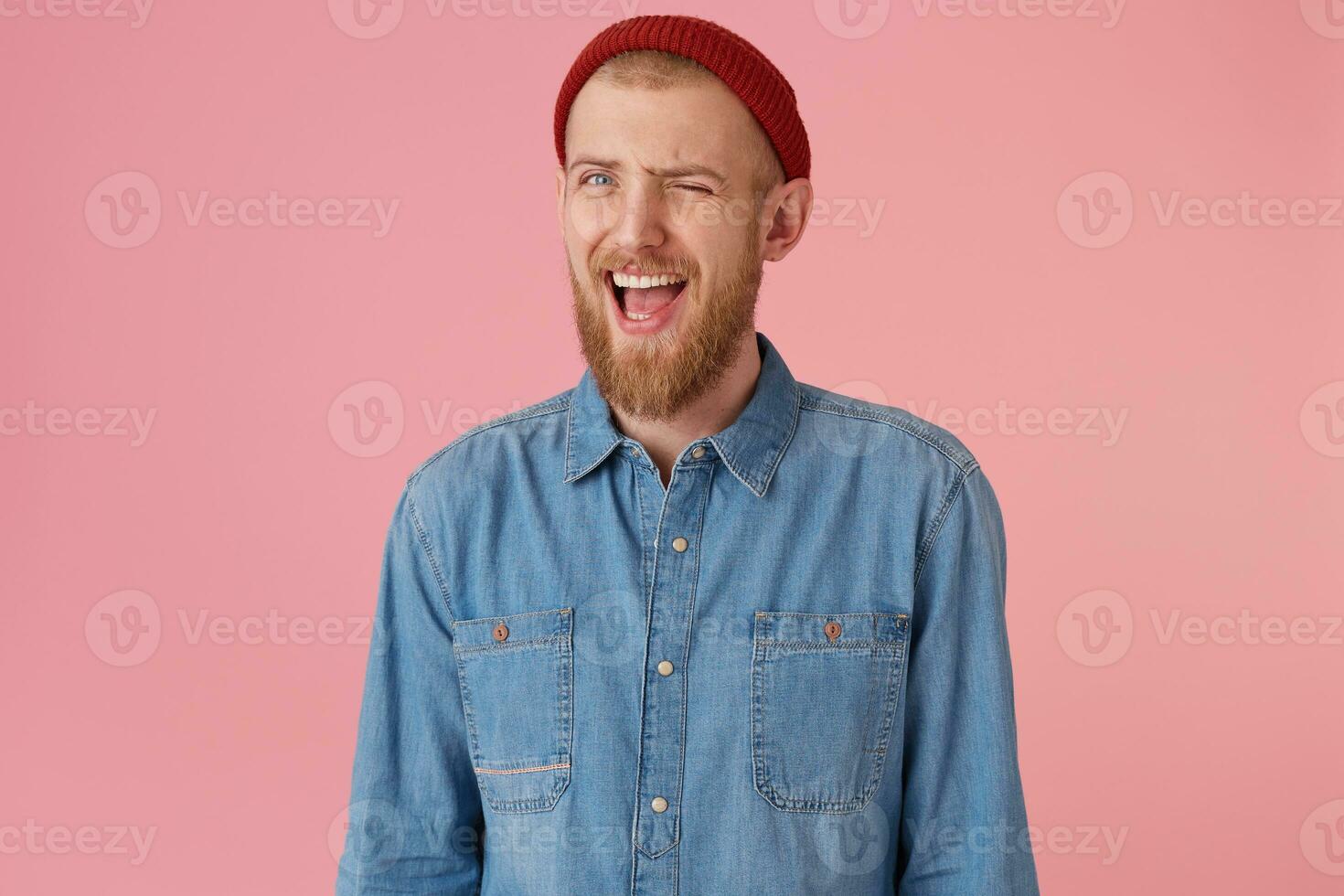 positif barbu Masculin avec façonné denim chemise rouge chapeau, a espiègle expression, clins d'oeil, bouche ouvert, encourage les soutiens, isolé plus de rose Contexte. Humain faciale expressions, corps Langue photo