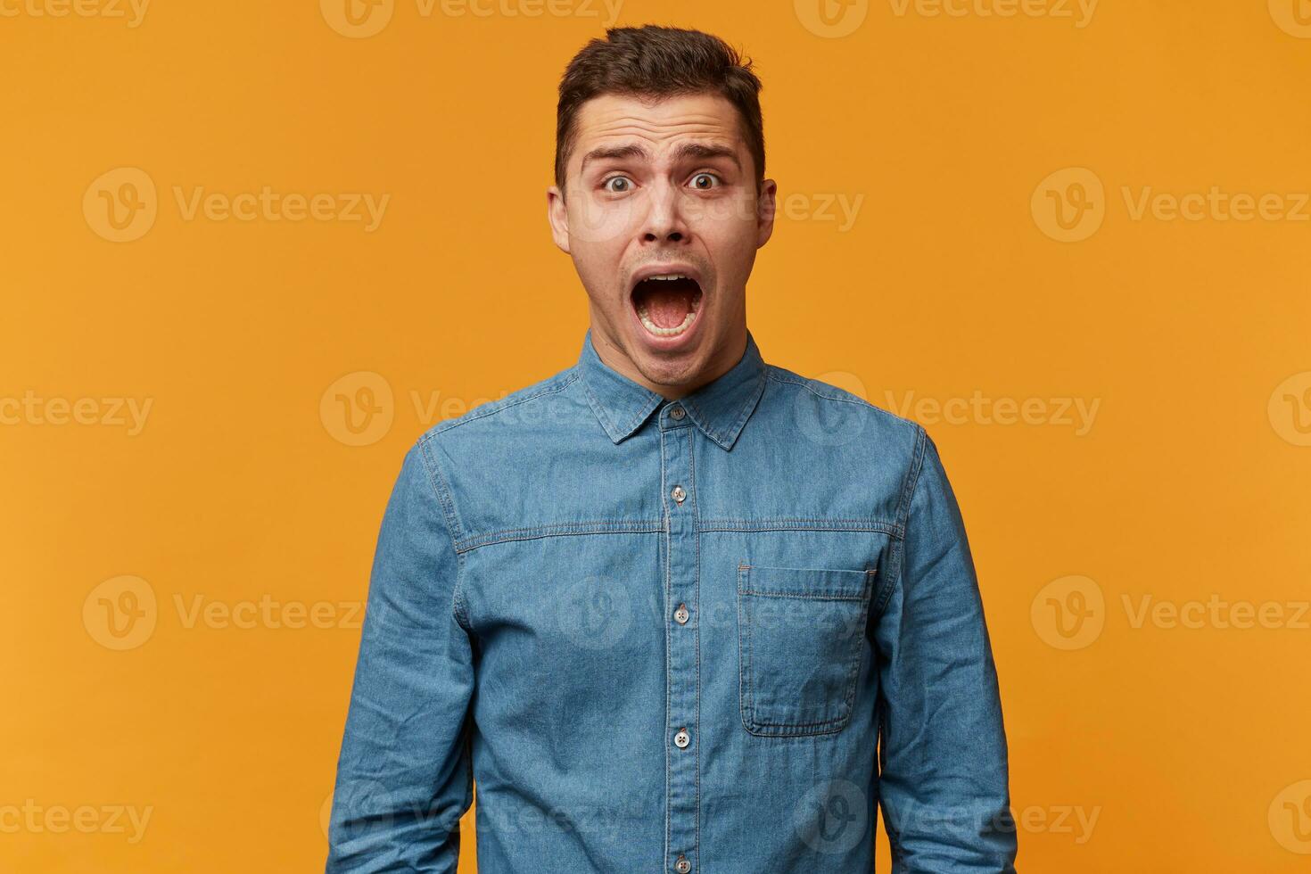 portrait de une gars qui cris dans une panique, en hurlant avec craindre, fait du une bruyant cri comme il est étonné et très effrayé, portant une à la mode denim chemise, isolé sur une Jaune Contexte photo