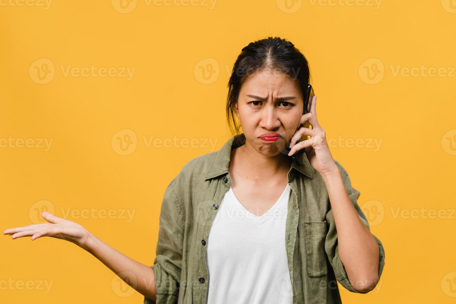 une jeune femme asiatique parle par téléphone avec une expression négative, des cris excités, des cris émotionnels en colère dans un tissu décontracté et se tient isolée sur fond jaune avec un espace de copie vierge. concept d'expression faciale. photo