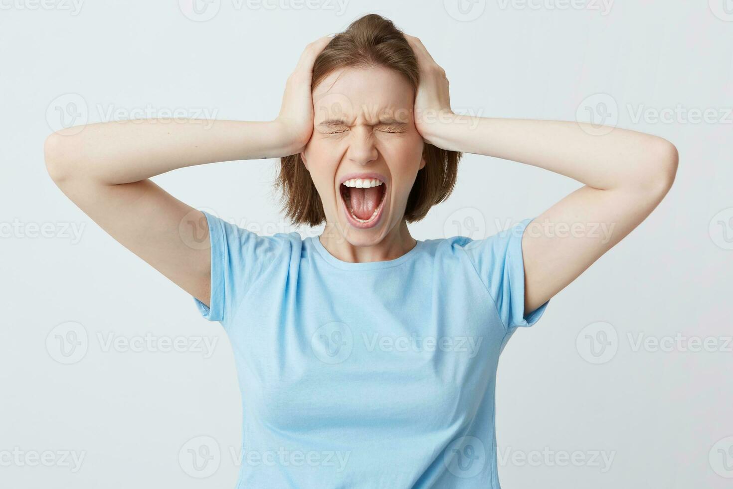 portrait de fou hystérique Jeune femme dans bleu t chemise avec fermé yeux et mains sur tête regards stressé et en hurlant isolé plus de blanc Contexte photo