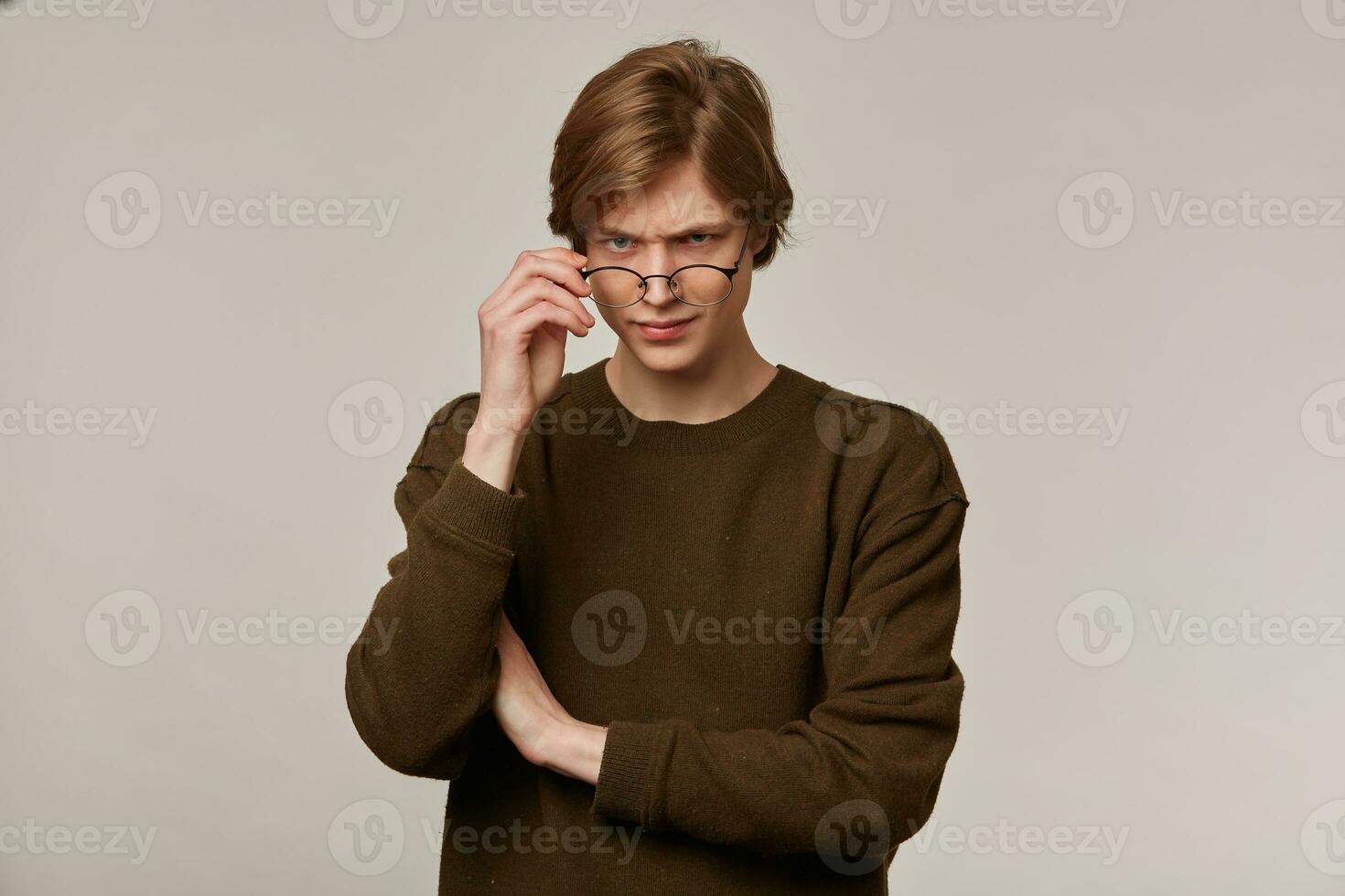 sérieux adolescent gars, incertain homme avec blond cheveux. portant marron chandail et lunettes. émouvant le sien lunettes. gens et émotion concept. en train de regarder soupçonner à le caméra isolé plus de gris Contexte photo