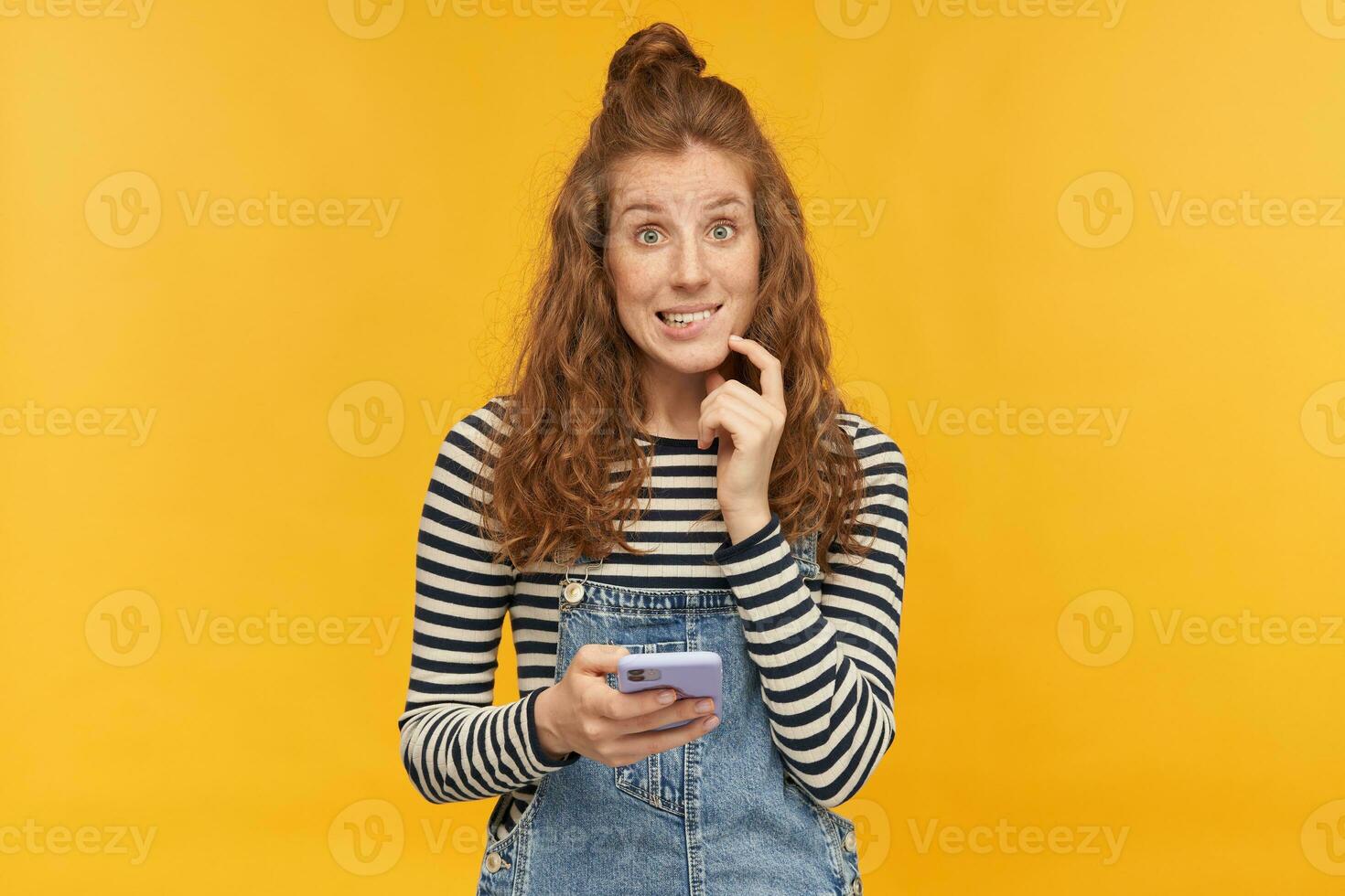 intérieur studio coup de Jeune gingembre femelle avec longue rouge frisé cheveux mordant sa lèvre, en portant téléphone avec honteux coupable faciale expression, isolé plus de Jaune Contexte photo