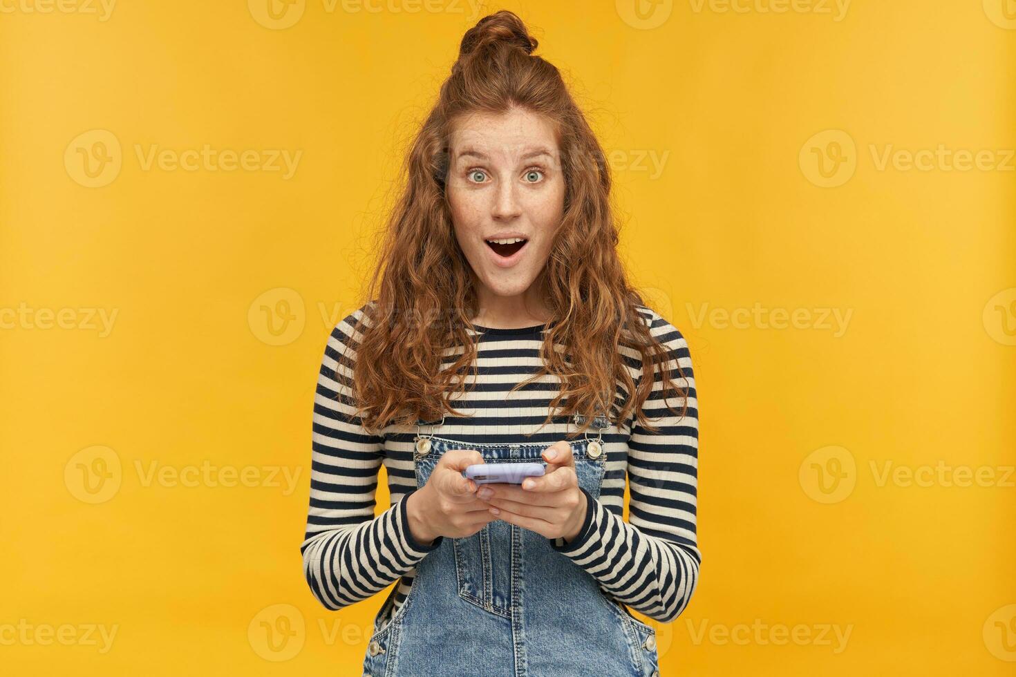 intérieur studio coup de Jeune gingembre femelle avec longue rouge frisé cheveux en portant sa téléphone regards directement dans caméra avec choqué, surpris faciale expression isolé plus de Jaune Contexte photo