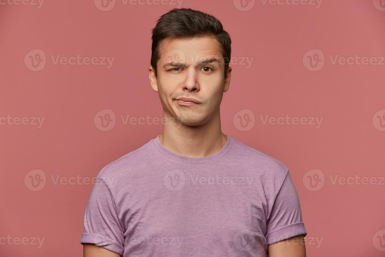 portrait de Beau renfrogné Jeune homme porte dans Vide T-shirt, regards à le caméra avec avec une sourire et les doutes, des stands plus de rose Contexte. photo