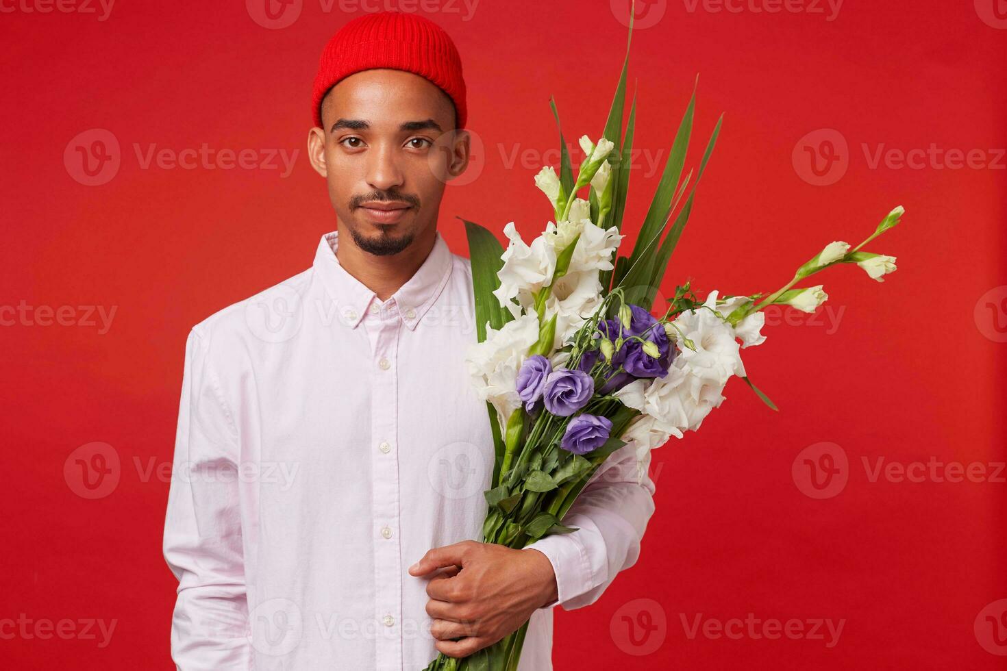 Jeune calme africain américain gars dans blanc chemise et rouge chapeau, détient bouquet , des stands plus de rouge Contexte et regards à le caméra. photo