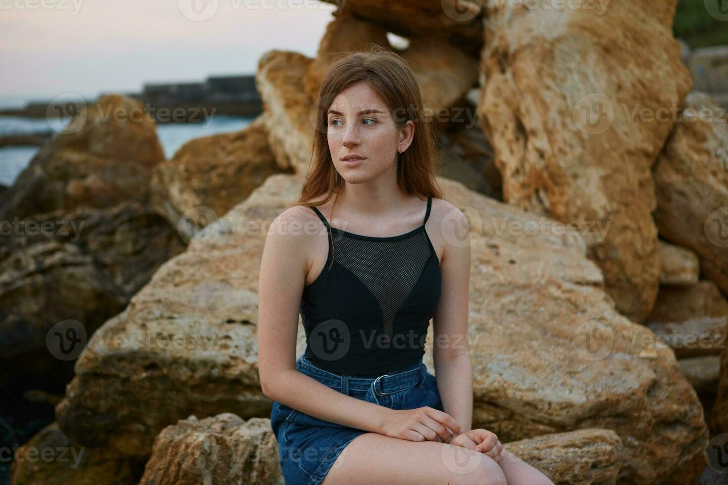 photo de Jeune roux mignonne Dame avec taches de rousseur est assis sur le plage, rêveusement regards une façon et sourire, regards positif et heureux.