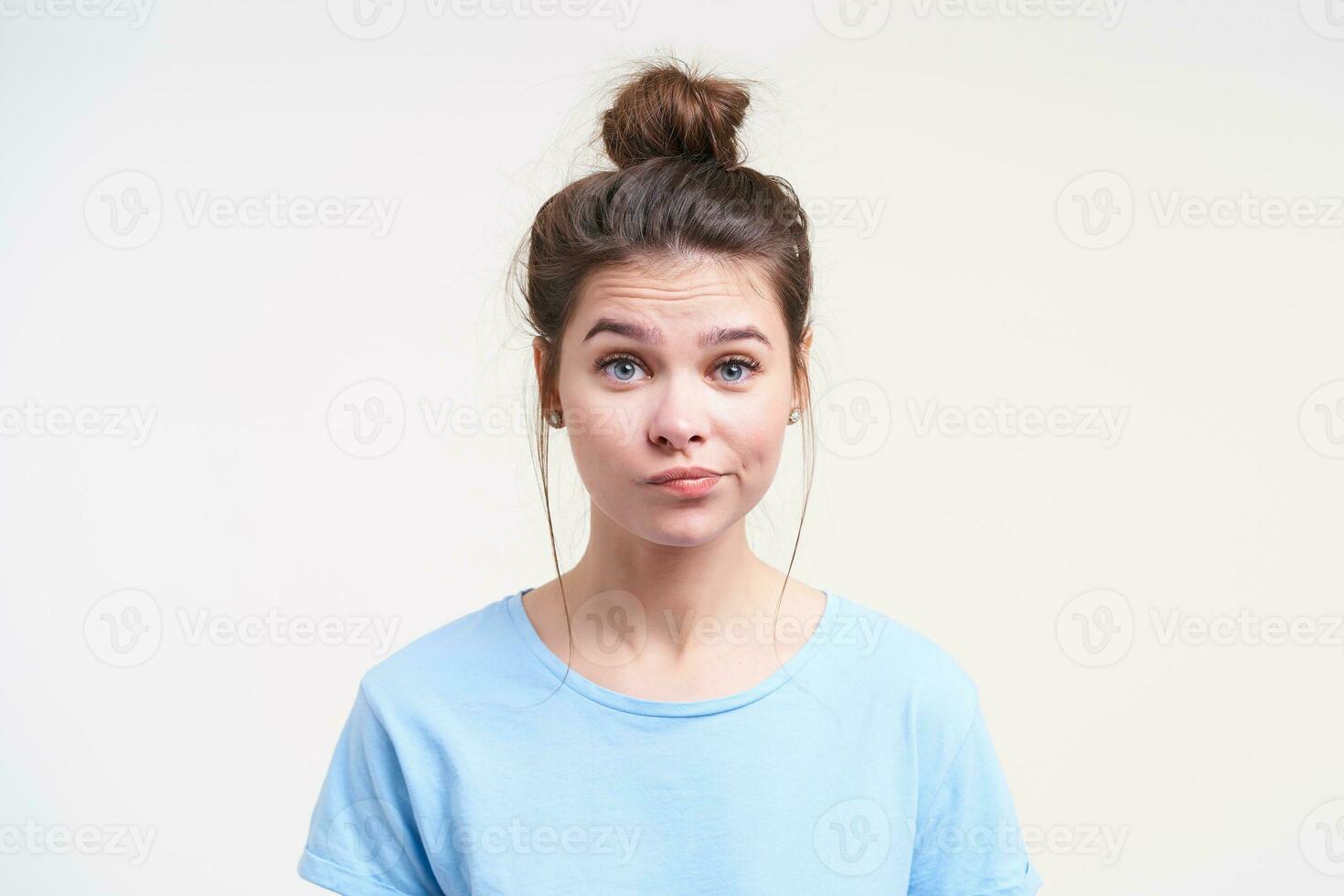 portrait de Jeune perplexe marron aux cheveux Dame avec chignon coiffure torsion sa bouche tandis que à la recherche avec perplexité à caméra, permanent plus de blanc Contexte photo