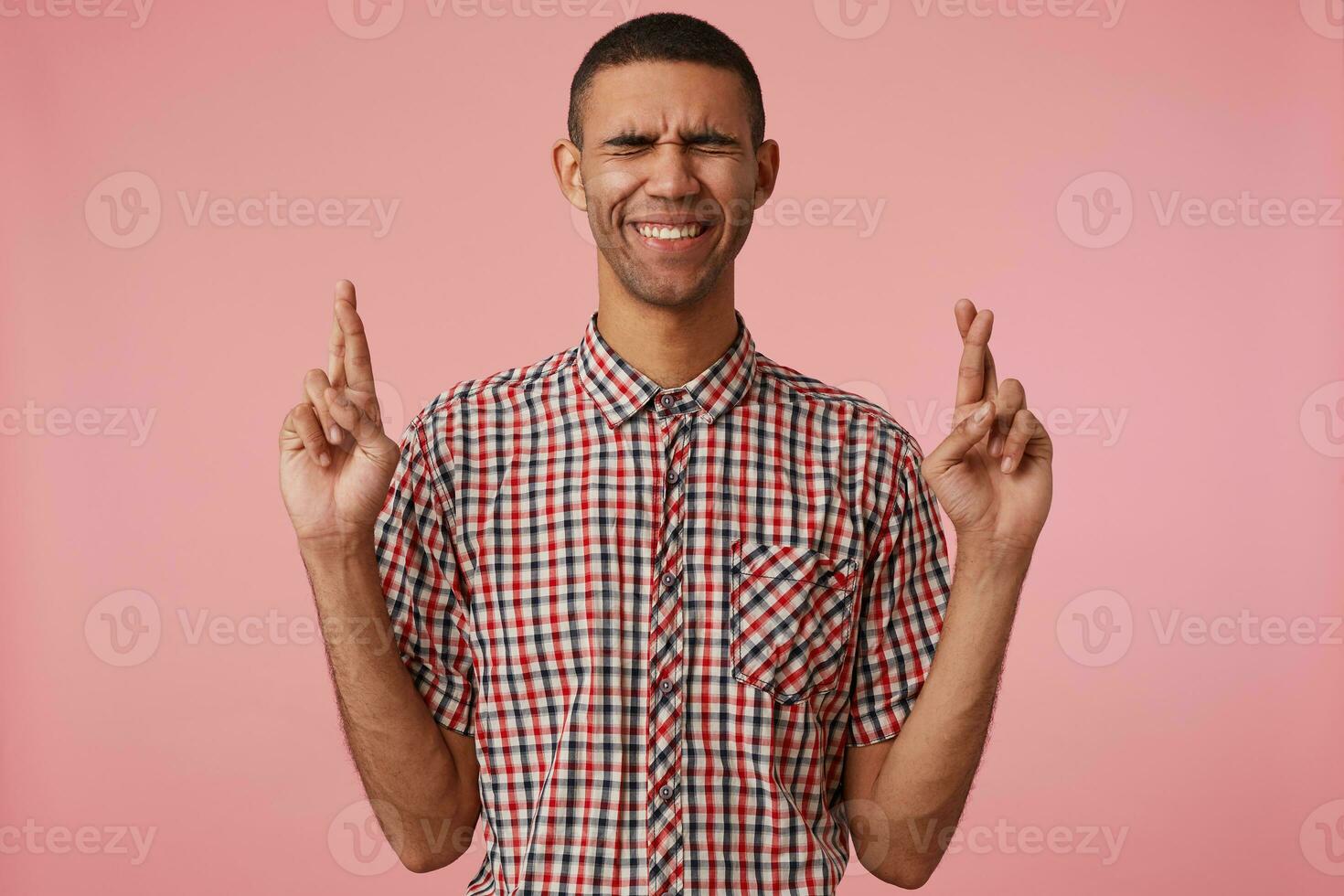 portrait de Jeune attrayant foncé écorché gars dans à carreaux chemise, avec franchi les doigts et fermé yeux, espère pour la chance et sourires, des stands plus de rose Contexte. photo