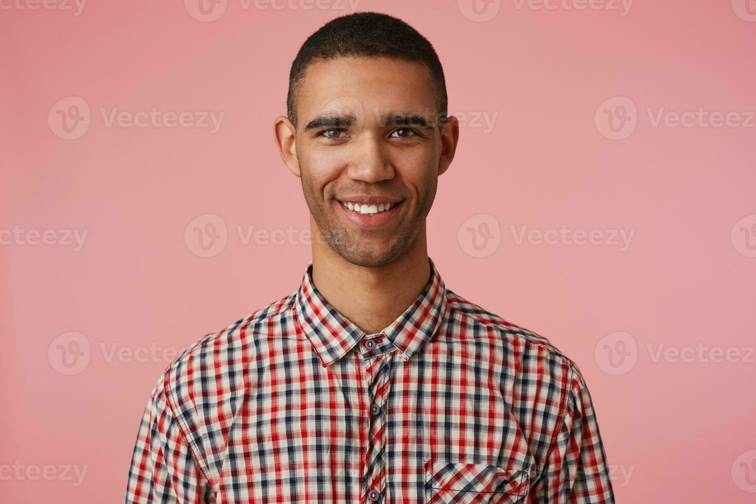 portrait de Jeune content attrayant foncé écorché gars dans à carreaux chemise, regards à le caméra avec positif expression, largement souriant et des stands plus de rose arrière-plan, positif émotion concept. photo