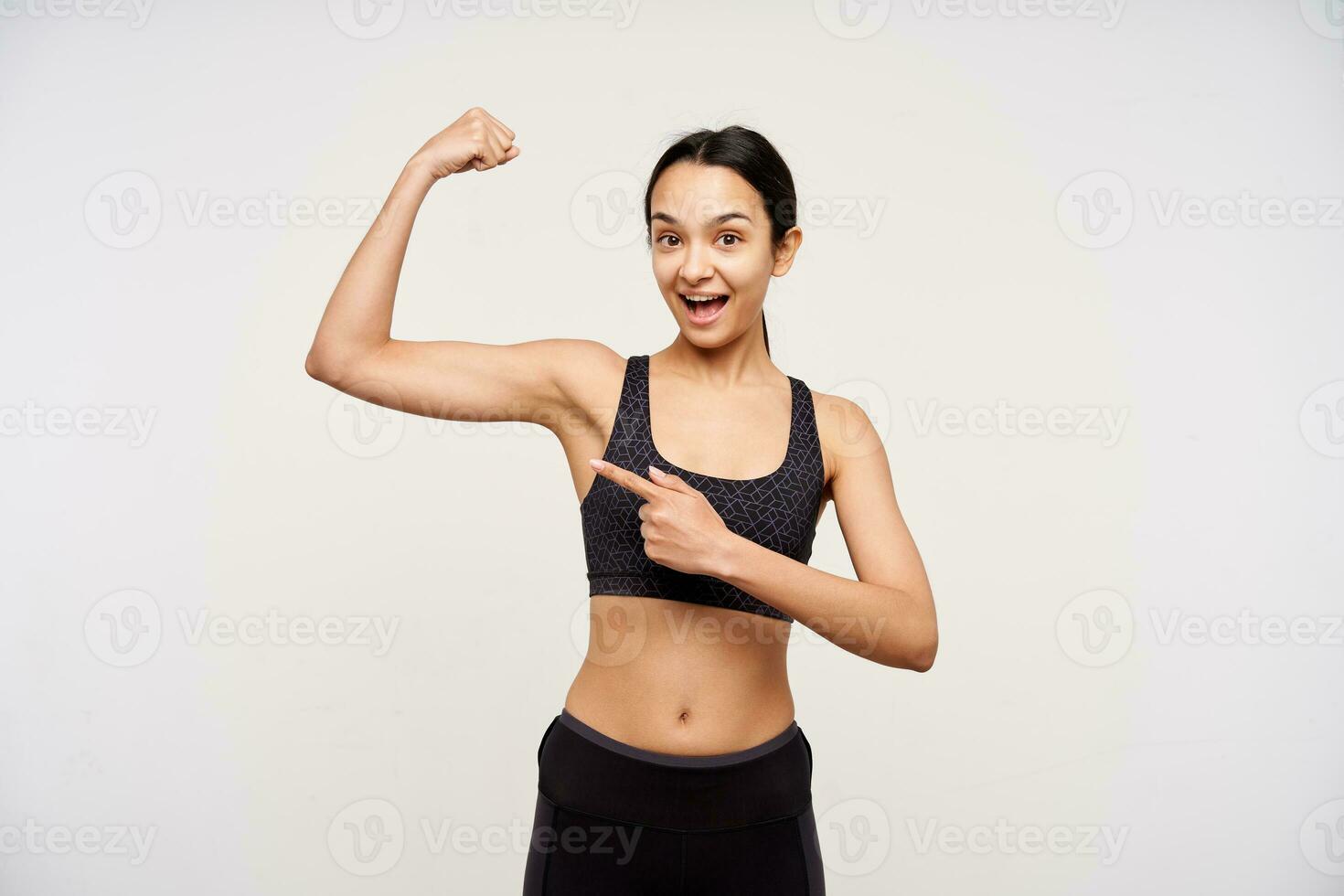 intérieur photo de Jeune sportif marron aux cheveux Dame avec décontractée coiffure montrer du doigt gaiement avec indice doigt sur sa élevé main tandis que permanent plus de blanc Contexte