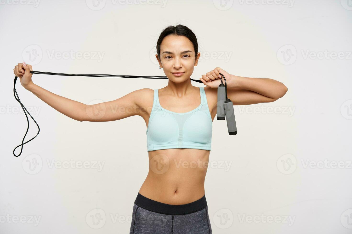 Jeune dame, sportif asiatique femme avec foncé longue cheveux. portant tenue de sport et élongation une sauter corde plus de sa cou. en train de regarder sur de soi à le caméra isolé plus de blanc Contexte photo