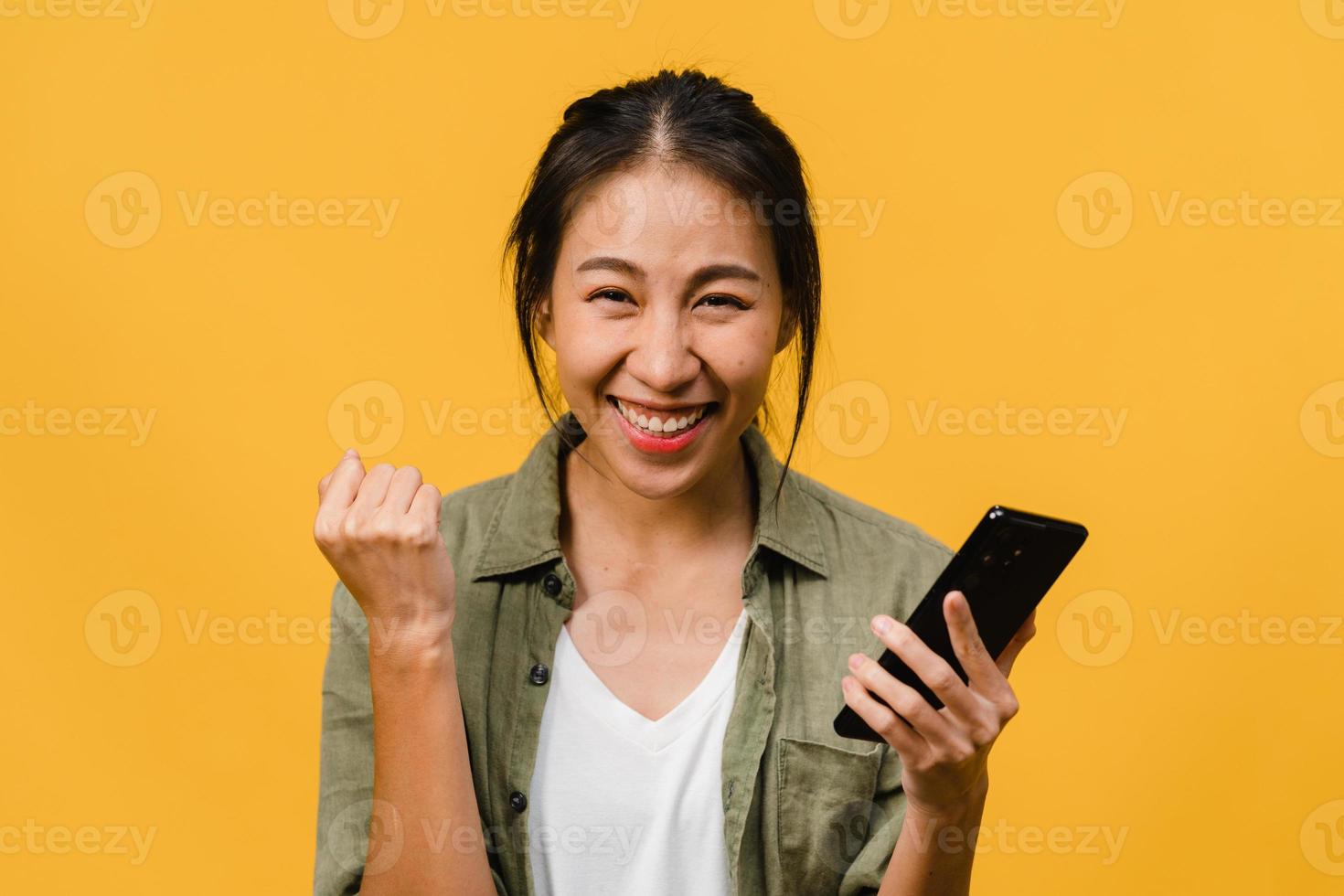 surpris une jeune femme asiatique utilisant un téléphone portable avec une expression positive, sourit largement, vêtue de vêtements décontractés et se tient isolée sur fond jaune. heureuse adorable femme heureuse se réjouit du succès. photo