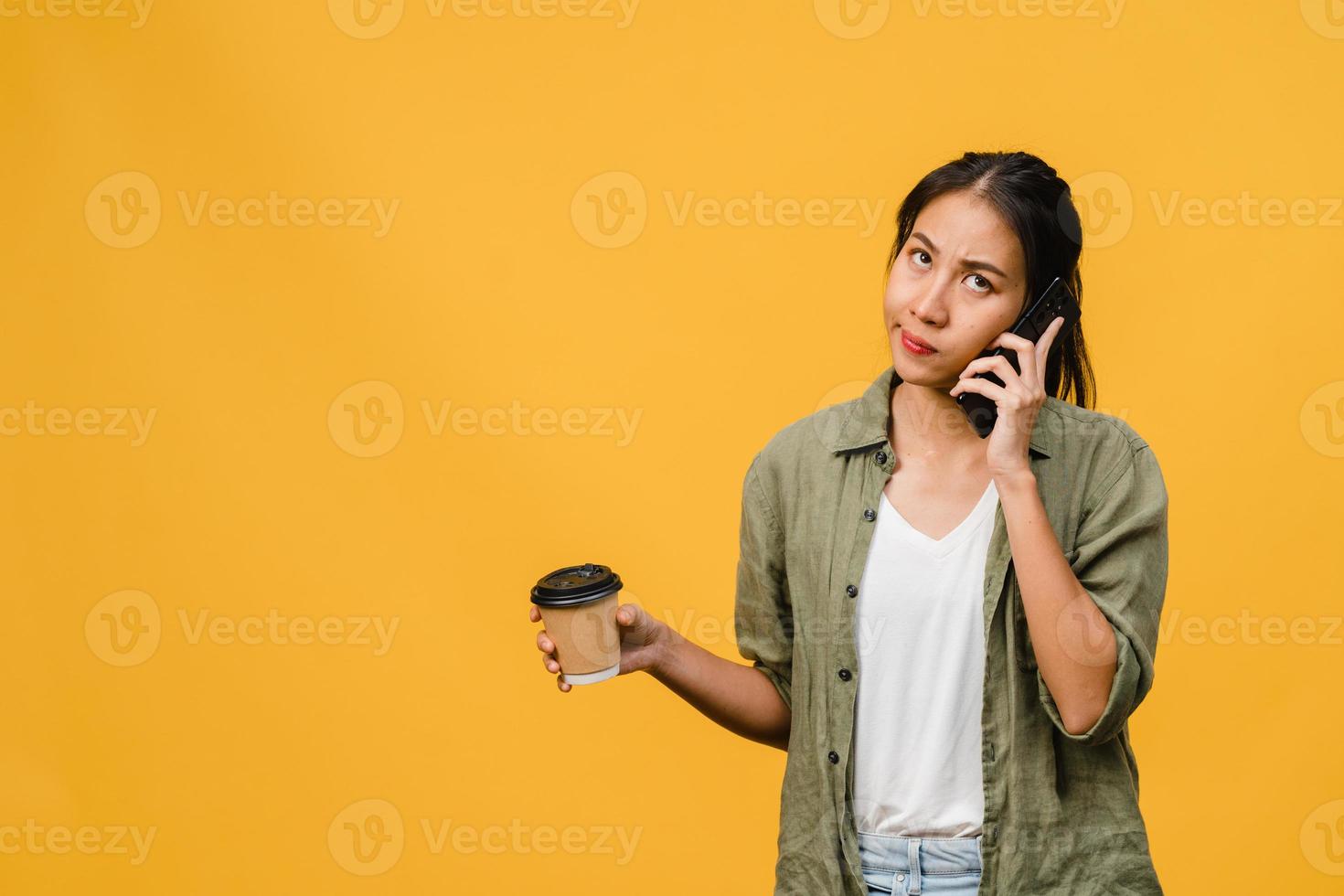 une jeune femme asiatique parle par téléphone et tient une tasse de café avec une expression négative, des cris excités, pleure émotionnellement en colère dans un tissu décontracté et se tient isolée sur fond jaune. concept d'expression faciale. photo