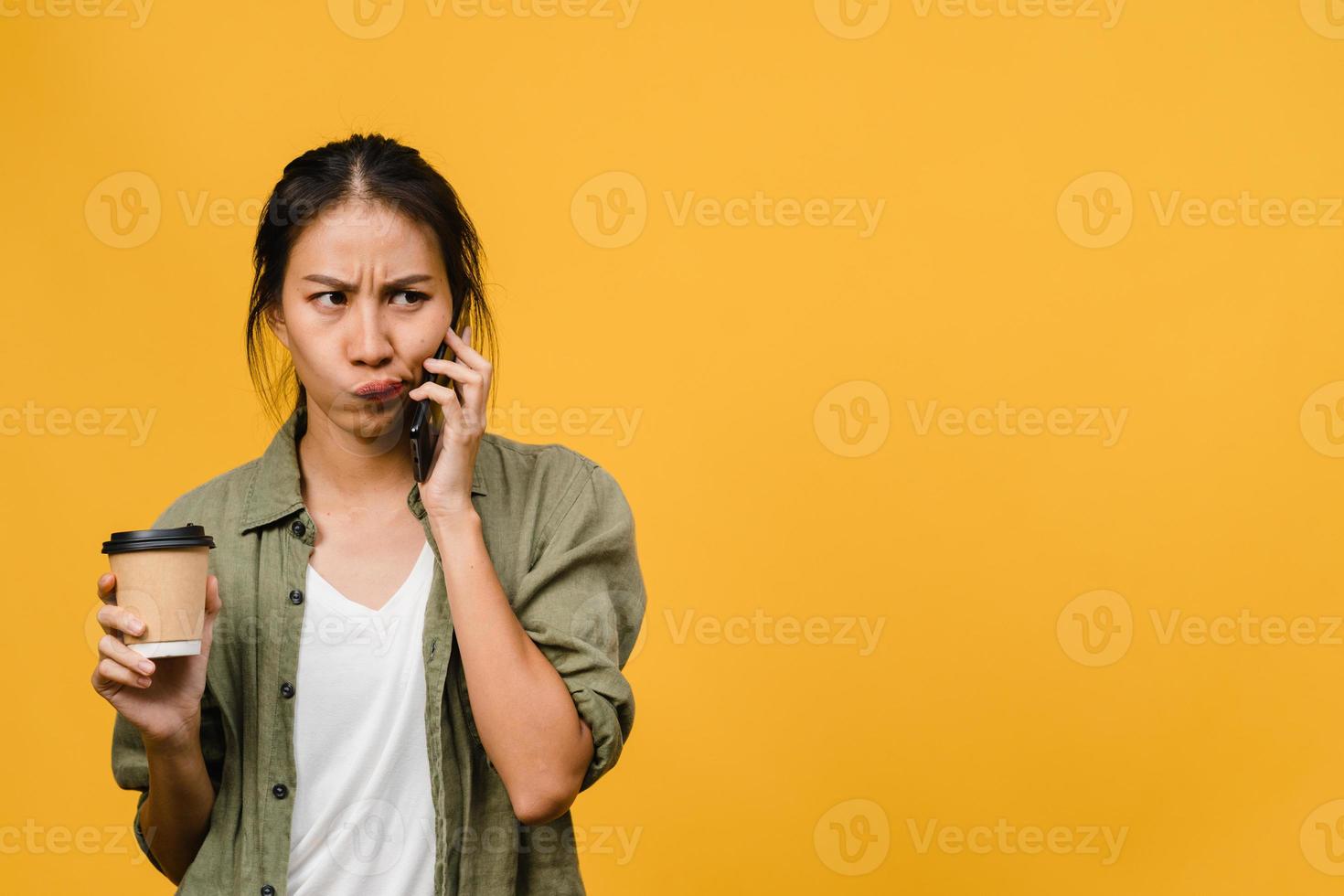 une jeune femme asiatique parle par téléphone et tient une tasse de café avec une expression négative, des cris excités, pleure émotionnellement en colère dans un tissu décontracté et se tient isolée sur fond jaune. concept d'expression faciale. photo