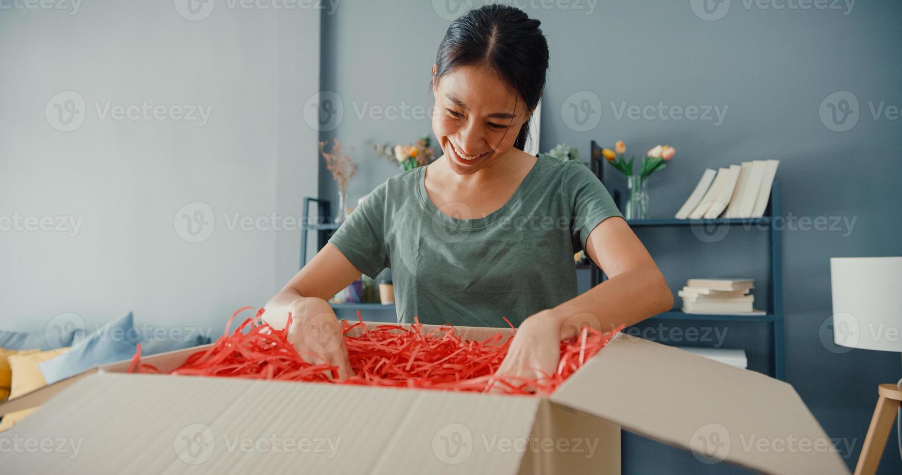 Heureuse belle dame asiatique déballant le colis de livraison en carton du marché en ligne dans le salon de la maison. acheteur satisfait dans les produits de déballage sur Internet, les achats en ligne et le concept de livraison. photo