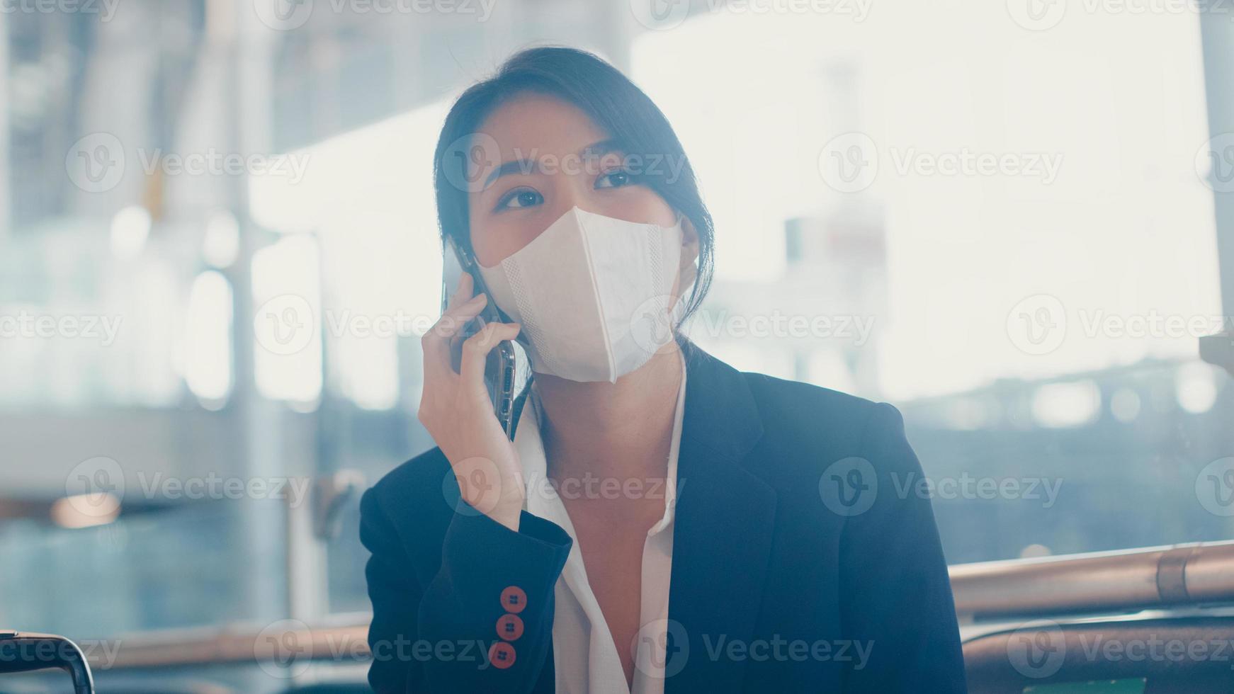 Une fille d'affaires asiatique intelligente porte un costume assis avec une valise sur un banc, appelez un téléphone intelligent avec un partenaire, attendez le vol à l'aéroport. navetteur de voyage d'affaires dans la pandémie de covid, concept de voyage d'affaires. photo